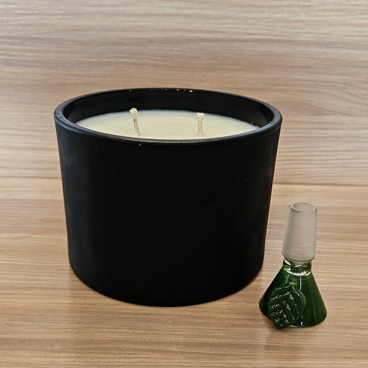 A black cylindrical candle jar with cream-colored wax sits on a light wood-grain surface next to a small green glass 14mm bowl piece. 