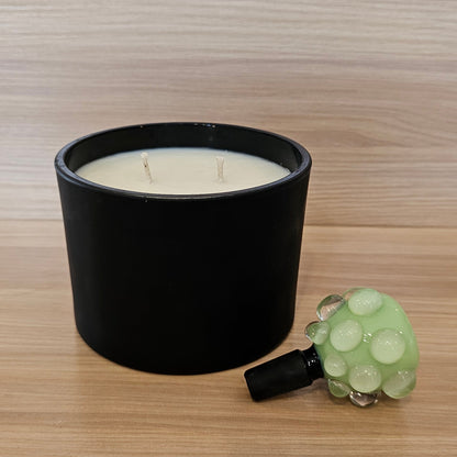 A black candle jar with white wax sits on a light wood surface next to a small green textured 14mm bowl piece, against a matching wood-grain background.