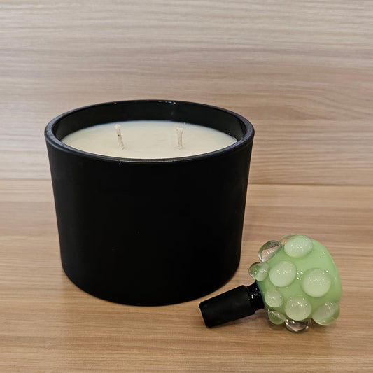 A black candle jar with white wax sits on a light wood surface next to a small green textured 14mm bowl piece, against a matching wood-grain background.