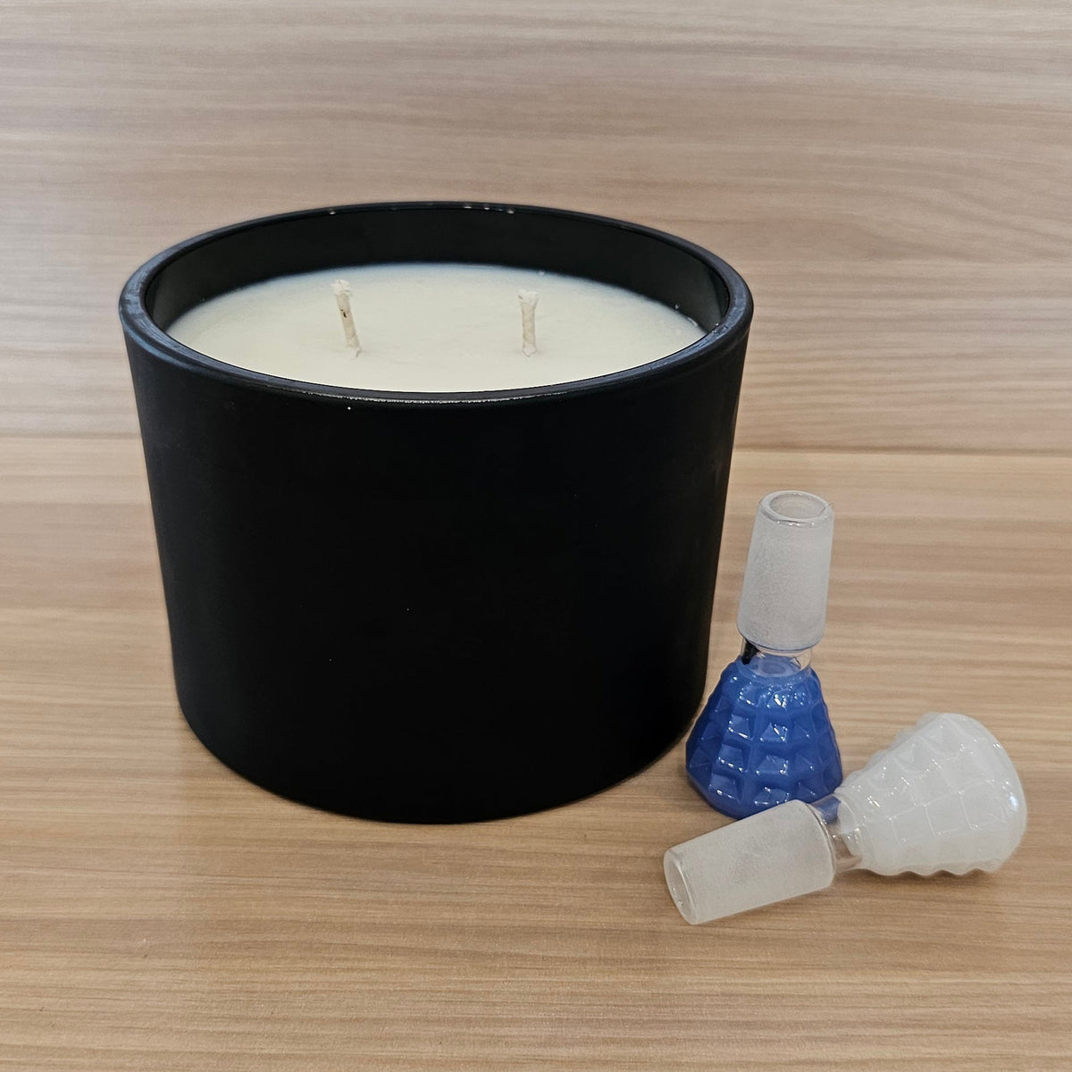 A black cylindrical candle jar with white wax sits on a light wood-grain surface, alongside a small 14mm bowl pieces. 