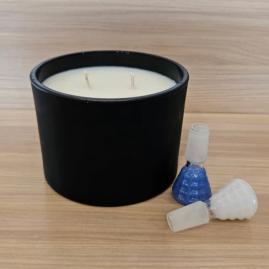 A black cylindrical candle jar with white wax sits on a light wood-grain surface, alongside a small 14mm bowl pieces. 