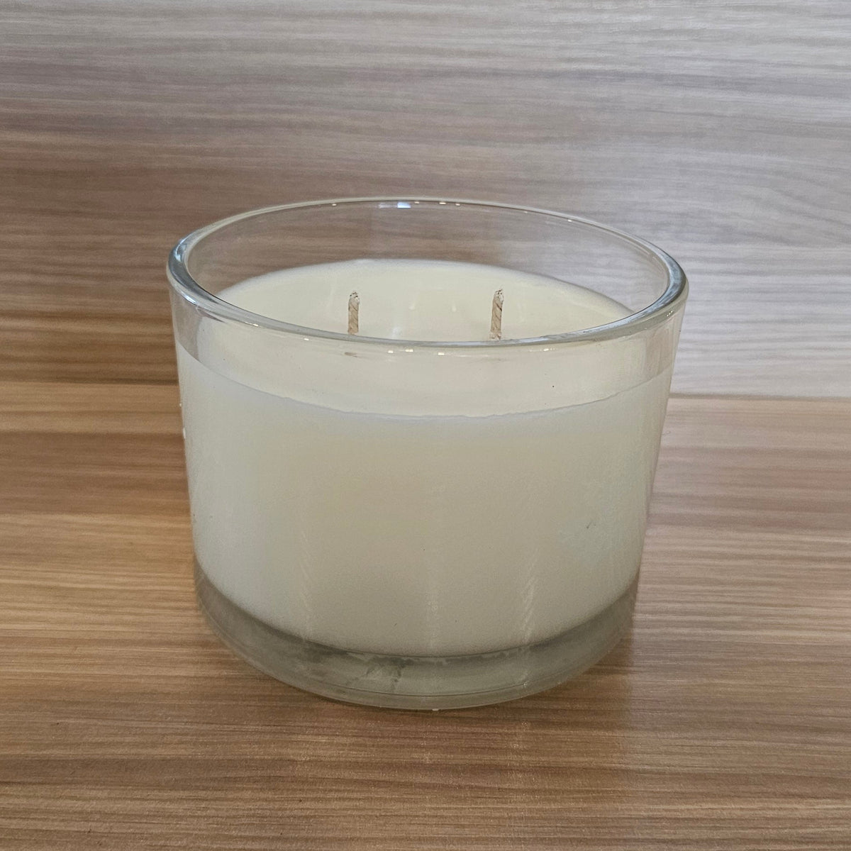 A double-wick soy candle in a clear glass jar, filled with off-white wax, set on a wooden surface against a wood-paneled background.