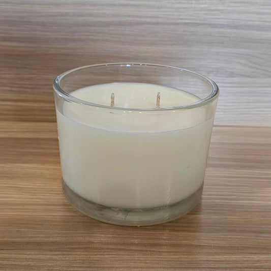 A double-wick soy candle in a clear glass jar, filled with off-white wax, set on a wooden surface against a wood-paneled background.