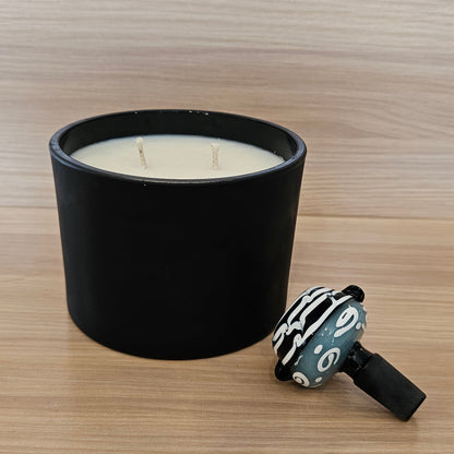 A black cylindrical candle jar filled with white wax sits on a light wood-grain surface next to a small black, white, and gray patterned 14mm bowl.
